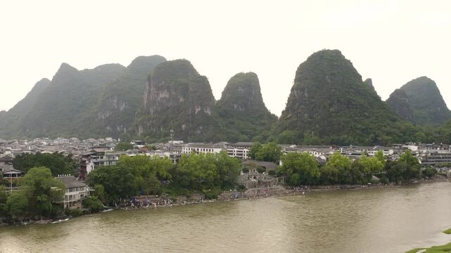 Upward drone ascent scales karst rampart above Yangshuo village, rooftops and river crowds shrinking as limestone peaks multiply across misty horizon in classic Guangxi landscape reveal.