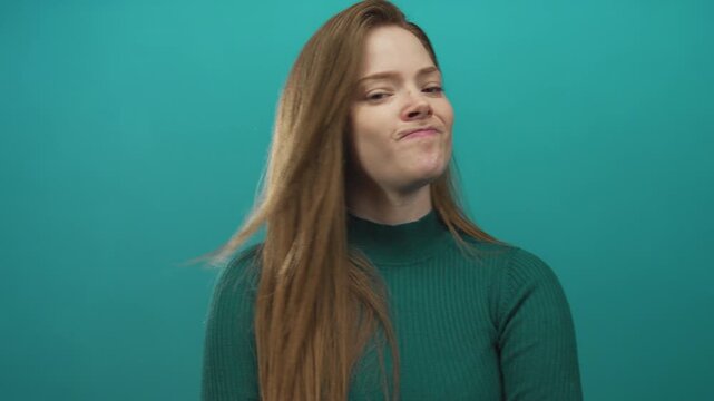 Woman folds arms and tilts head sideways with mouth open in studio under soft lighting framed by teal backdrop; denial.