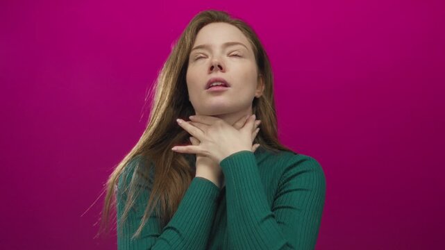 Young caucasian woman holds hand on neck for pain with grimacing expression against a pink studio wall; discomfort.