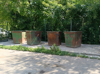 Naklejka premium Row of heavily rusted industrial trash bins sits in the bright sun, surrounded by lush green trees and a chain-link fence. Waste disposal
