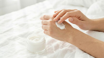 A person gently applies white moisturizing cream from a glass jar to their hand, emphasizing daily skincare and wellness on soft white bedding.