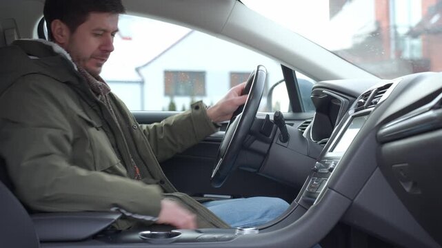 Freezing man dressed in jacket sitting in parked car and and pressing climate control button on dashboard during cold winter day