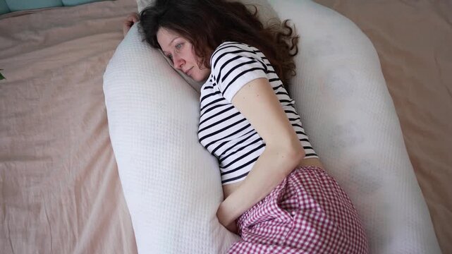 Overhead view of a pregnant woman in her pajamas waking up in bed, lying on her side and surrounded by a large white u-shaped maternity pillow for ultimate comfort during her sleep