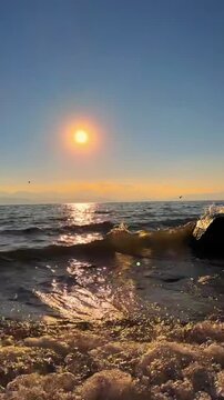 CHINA: 1080p vertical video (9:16). A cinematic low-angle close-up shot of water waves rolling and crashing onto the shore. The sun is setting in the background, casting a bright golden glare and shim