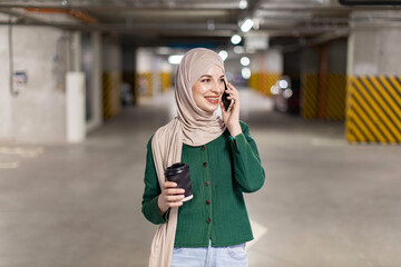 A smiling woman wearing a hijab talks on her phone while holding a coffee cup in a parking garage