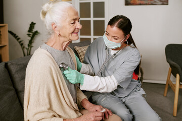 Obraz premium Senior Caucasian woman sitting on sofa receiving medical examination from young adult Asian female nurse wearing scrubs and mask using stethoscope in home setting