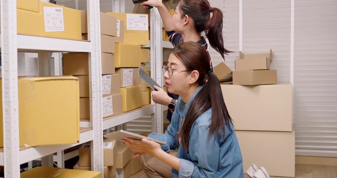 Asian women online business partners scanning parcel box label, cross checking stock quantity on tablet, organizing online shopping inventory inside home e-commerce warehouse shelf