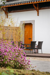 Traditional Zen Style Courtyard with White Wall, Wooden Door, Bamboo and Purple Flowers in the Foreground. Peaceful Outdoor Patio with Wicker Chairs and Table in a Serene Garden Setting.