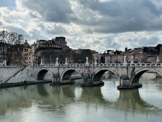 Obraz premium Wide-angle view of Ponte Sant’Angelo in Rome from a distance, with the Tiber River below and angel statues lining the historic bridge against the city skyline