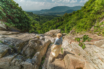 Young woman in straw hat standing on rocky waterfall edge overlooking lush green valley and mountains on Koh Samui Thailand enjoying tropical landscape and travel adventure