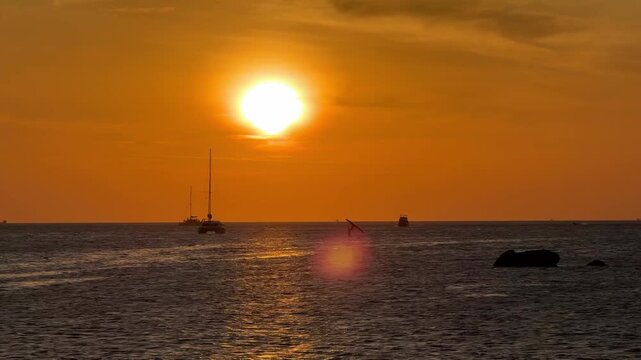 blazing red setting sun sinking into the Andaman Sea near southern Phuket, Thailand. Fiery sunset colors fill the sky as a distant anchored yacht appears in dark silhouette on the horizon