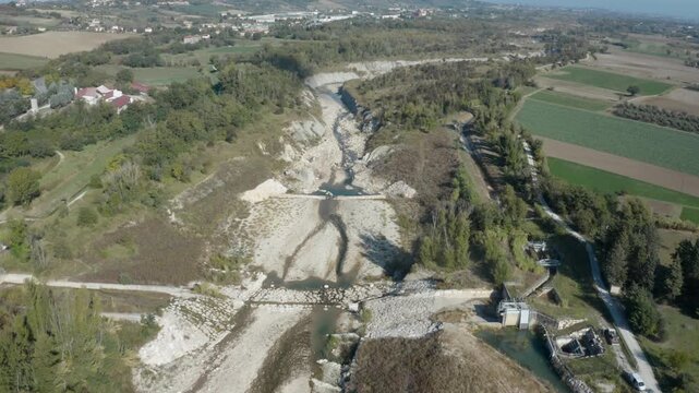 Drone view of a dry riverbed with levees and control gates, surrounded by farmland, trees, and rural roads.