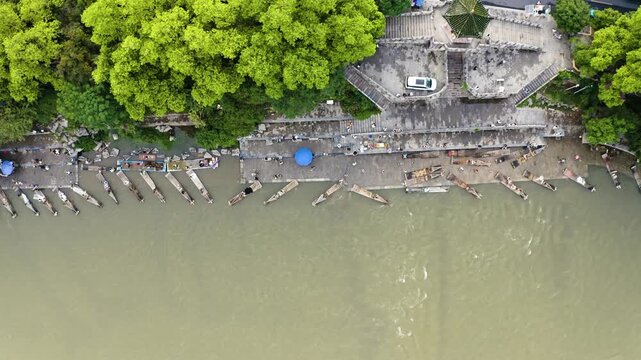 Top-down descent plunges from lofty heights to verdant riverbank wharf, bamboo rafts aligned under leafy bowers, figures animating stone piers against serene Li River flow.