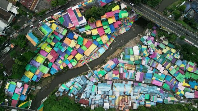 Overhead drone view of a colorful urban village with painted houses, bright rooftops, structured neighborhood layout and a bridge spanning a winding river in Malang City Indonesia