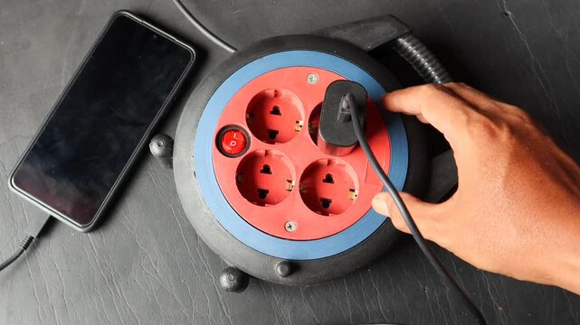 Asian man hand plugging a power adapter into an extension socket beside a smartphone.
