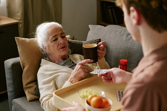 Senior Caucasian woman reclining on sofa holding mug, while young adult Caucasian woman offering medication and tray with fruit slices and glass of water during home care