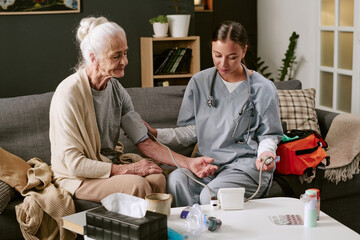 Obraz premium Senior Caucasian woman sitting on sofa receiving blood pressure measurement from young adult Caucasian female nurse using medical device, healthcare equipment visible on table