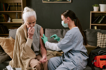 Senior Caucasian woman sitting on sofa receiving vaccination from young adult Caucasian female...