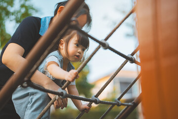 Happy little Asian girl climbing a rope net at an outdoor playground with her mother supporting...