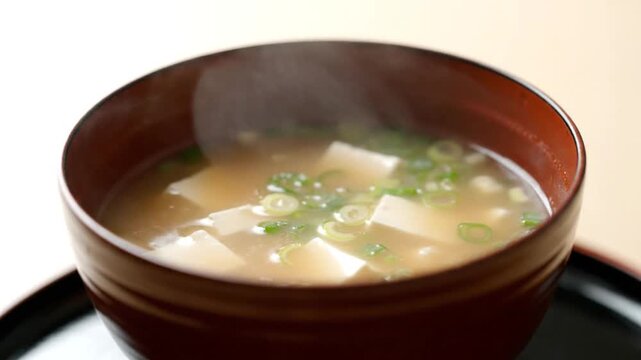 Steaming bowl of miso soup with tofu and green onions on a black tray against a white background.