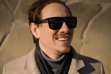 Obraz premium Close-up portrait of a young caucasian redhead man with sunglasses smiling wearing a coat on a winter day while walking down the street. Lifestyle.