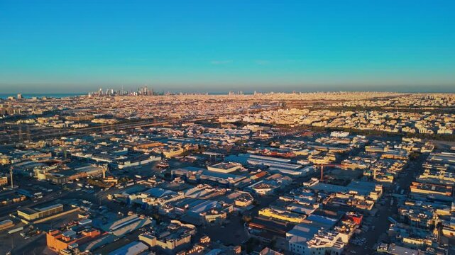 Wide drone panorama of Kuwait City in golden hour light, showing sprawling neighborhoods, industrial rooftops, long highways and distant downtown skyline near the Arabian Gulf coastline.