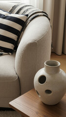 A close-up view of a textured cream armchair with a black and white striped pillow and a unique sculptural ceramic vase featuring multiple small openings.