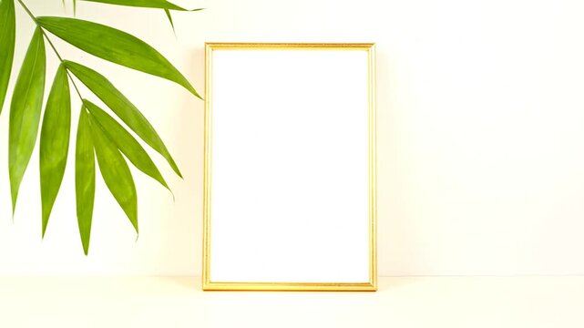 A gold framed rectangle blank canvas rests on a beige surface with green leaves in the upper left corner