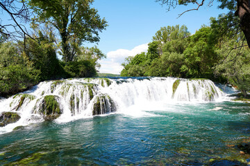 Fototapeta premium Majestic waterfall cascades through lush green nature. Clear turquoise water flows under a blue sky, conveying serenity and natural beauty