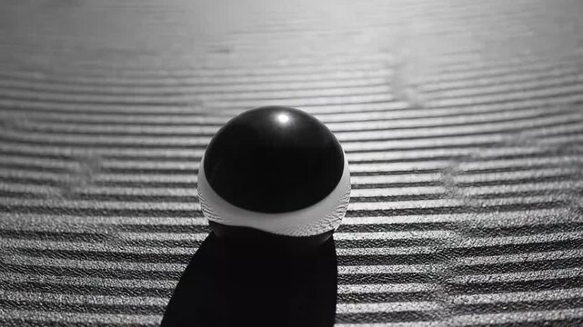 Abstract still life featuring a reflective. Half black half white sphere dripping a dark fluid onto a subtly textured. Raked surface against a plain black background. Representing contrast and duality