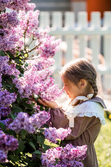 Vintage women's accessories and retro-style clothing. A beautiful little girl with bows in her hair, wearing a beige dress and lilac flowers in the open air.