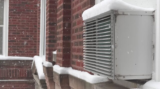 Snow is falling on a window air conditioner unit on a brick building. Close-up of a cold winter day in Chicago. Urban winter weather concept.