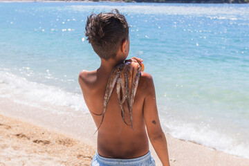 Boy on beach holding fresh octopus over his shoulder facing the turquoise sea