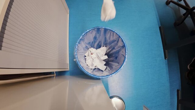 Overhead office shot of a wastebasket as a crumpled sheet of paper is tossed in, capturing a simple daily workplace moment. Ideal for concepts like rejected ideas, mistakes, cleanup, productivity, and