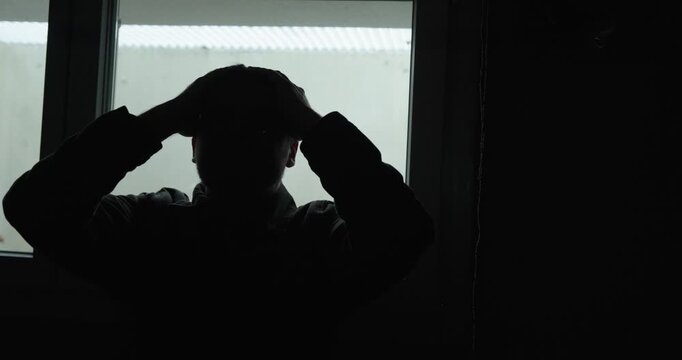 Cinematic silhouette of a man sitting near a bright window, holding his head in distress and deep sadness. Ideal for concepts like depression, anxiety, burnout, loneliness, emotional crisis, and menta