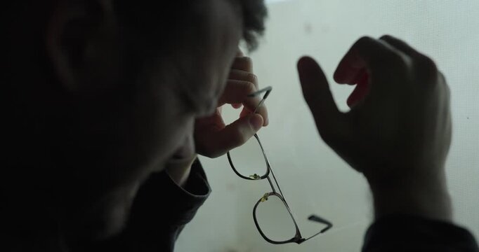 Emotional close-up of a man near a window wiping tears while holding his glasses, conveying grief and exhaustion in a dark, cinematic mood. Ideal for mental health, depression, burnout, loss, and pers