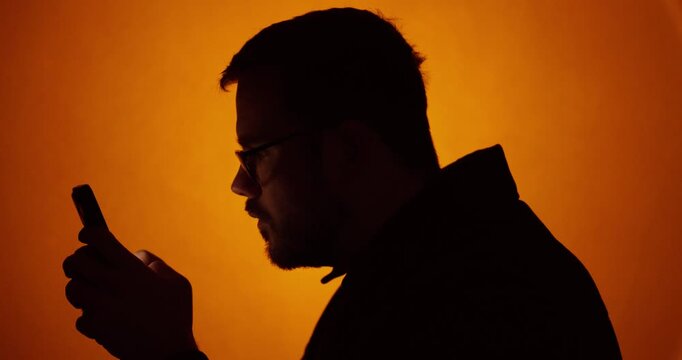 Tight side-profile silhouette of a man focused on his smartphone against a warm orange background, creating a cinematic and anonymous tech mood. Ideal for messaging, digital addiction, privacy concern