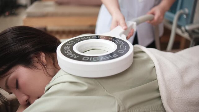 Asian female patient's back is treated by physical therapist for pain relief with PMS shockwave device, muscle recovery therapy at wellness healthcare spa in doctor's clinic of orthopedic hospital.