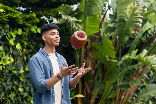 Adult man tossing American football while wearing denim shirt and bracelets in backyard garden