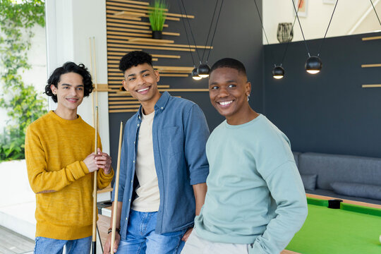 Diverse male friends posing and smiling beside green-felt pool table in modern lounge holding cues