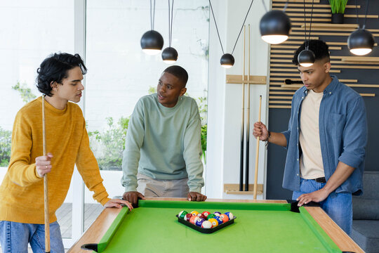 Diverse male friends leaning around felt pool table in rec room holding cues watching racked balls