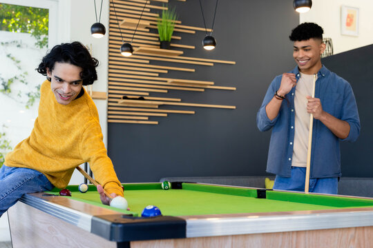 Male friends playing pool in modern recreation room, one leaning and aiming at green-felt table