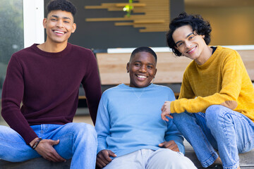 Three male friends sitting on steps in modern building with wooden panel wearing sweaters and jeans