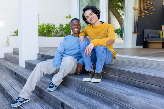 Diverse male partners sitting on wooden deck steps smiling, one resting arm, sunglasses perched