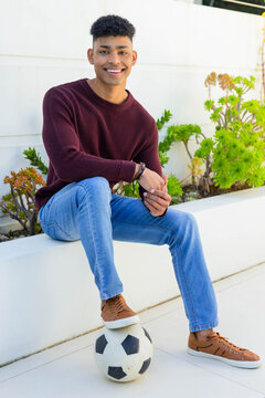African American man sitting on planter in courtyard in maroon sweater, resting foot on soccer ball