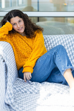 Woman in her twenties reclining on houndstooth sofa in living room, wearing mustard-yellow sweater
