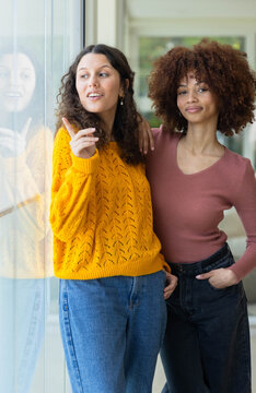 Diverse female friends posing in hallway pointing by glass window wearing sweaters and jeans