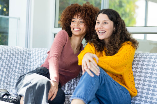 Two female friends sitting on patterned sofa in living room, smiling and wearing bright sweaters