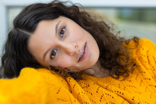 Female reclining toward camera at home, holding eye contact, wearing mustard-yellow sweater
