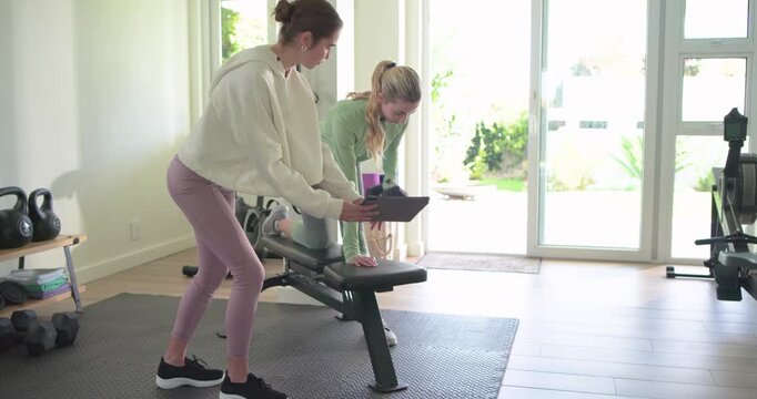 Female trainer adjusting tablet while guiding client rowing dumbbell on bench in gym for form